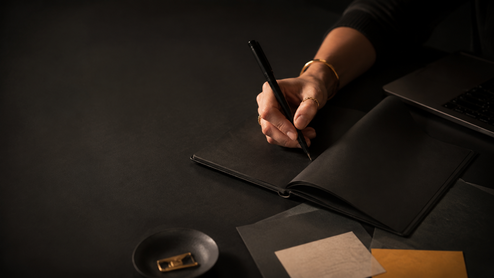 Hand writing in a notebook beside a laptop on a dark desk